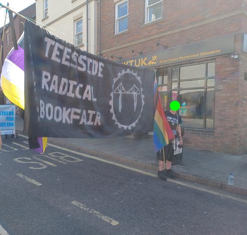 same as previous photo but taken from the other side of the street. non-binary flag at the left of banner, pride flag on the right.