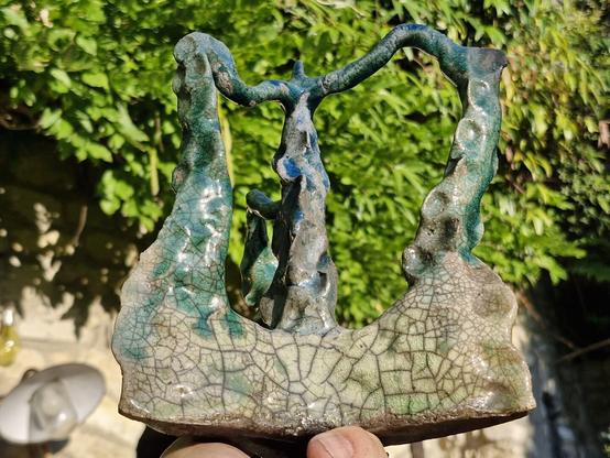 Sur un fond de végétation floue, une main tient un objet en terre cuite recouvert d'émaux vert, belu et blanc-gris. Des sortes de filaments ou de tentacules se rejoignent par leurs extrémités, qui semblent émerger d'une base triangulaire.