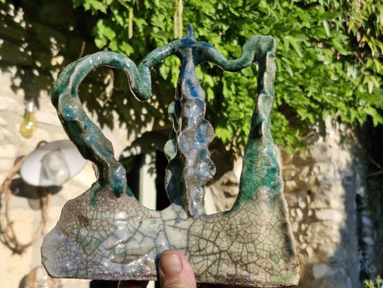 Sur un fond de végétation floue, une main tient un objet en terre cuite recouvert d'émaux vert, belu et blanc-gris. Des sortes de filaments ou de tentacules se rejoignent par leurs extrémités, qui semblent émerger d'une base triangulaire.