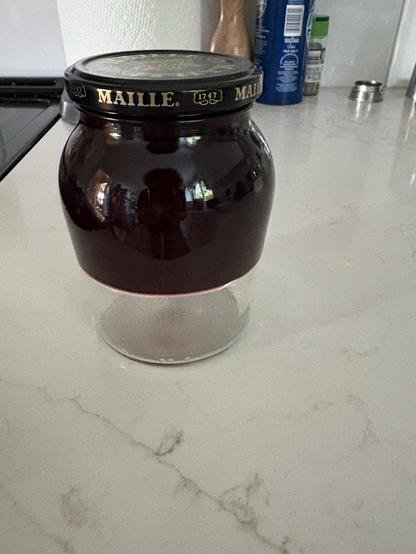 A glass jar on a white counter. There top two thirds is the jar are filled with dark red jelly, the bottom third is empty
