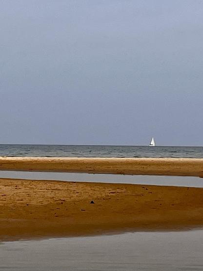 A serene coastal scene featuring a sandy beach with a shallow tidal pool in the foreground and calm Lake Michigan waters in the background. A small white sailboat is visible on the horizon under a cloudy sky.