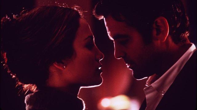 The image shows a close-up of two individuals facing each other, captured in profile against a warm, softly lit background. On the left, a woman with her hair pulled back into a bun and wearing a dark top gazes intently at the man on the right. The man, with short hair, is dressed in a white collared shirt and has a thoughtful expression. The background exhibits blurred, circular lights, enhancing the intimate atmosphere with a reddish hue.