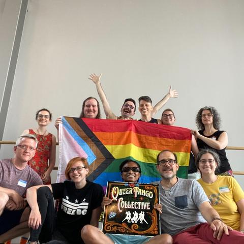 11 people gather around a Pride flag. The back row stands, holding it, except for that one person whose arms are raised. The front row sits with the queer tango collective” sign.