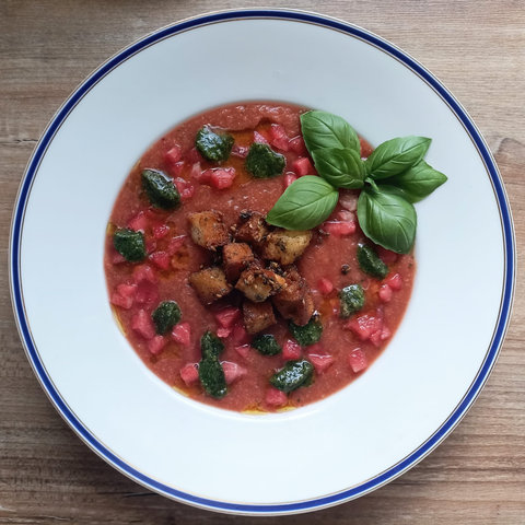 Ein Teller mit roter Suppe, Melonenwürfeln, Brotwürfeln + Pesto dekoriert mit einem Zweig Basilikum.