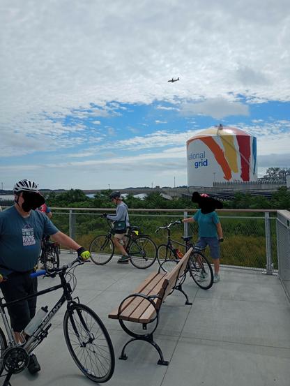 Park bench, people standing next to bicycles. Large gas tank in the background with the rainbow swish mural
