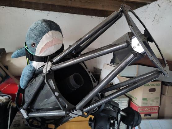 A Blåhaj cuddly shark sitting on an open mirror telescope, looking from the side at the mirrors. The Blåhaj wears a haj-vis harness with rainbow patches.

It is a Cassegrain Reflector Telescope, Hapi, a bit like the biggest telescopes in the world!
