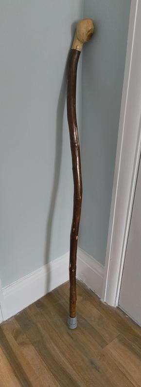 A walking stick made of stained wood - it is certainly not straight.
It has a heavy light wood handle and a grey ferrule.
It's in the corner of two light blue walls on a wooden floor.