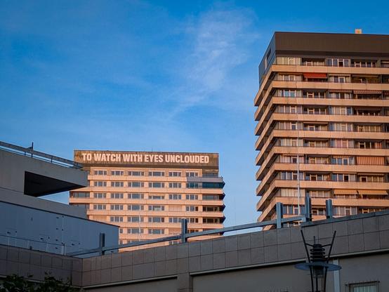 Color photo of two tall residential buildings at sunset, with a clear blue sky in the background. On the facade of the top floor of one building, there is a large inscription that reads “TO WATCH WITH EYES UNCLOUDED.”