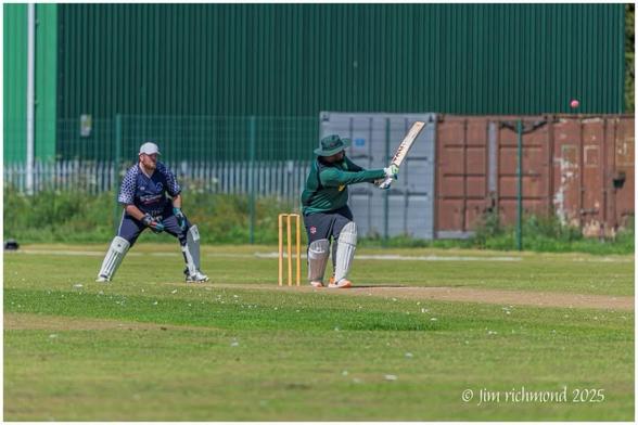 A cricket match scene featuring a player in green batting, attempting to hit a pink ball. Another player in blue and gray is positioned as the wicketkeeper behind the stumps. The background shows a green metal structure and some storage containers. Bright sunny weather.