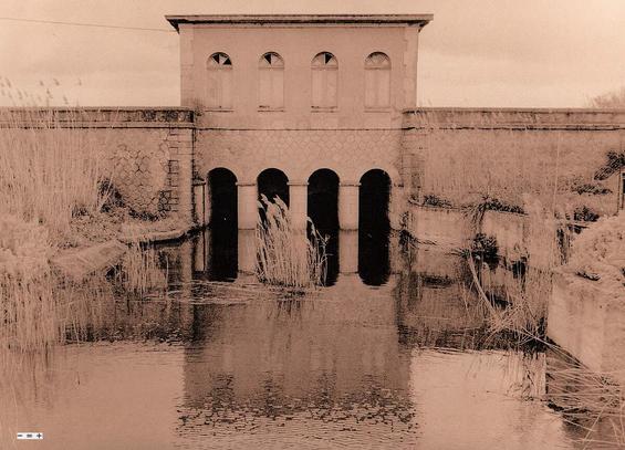 Ein steinernes Wasserwerk auf Sardinien mit vier Bögen, durch die Wasser fließen kann. Davor ein mit Schilf bewachsenes Becken. Die Anlage sieht aus wie seit Jahren außer Betrieb. Das monochrome Foto ist ein Scan von einem Handabzug aus der Dunkelkammer im Lithverfahren.