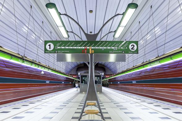 Symmetrical view on colourful metro platform at Petriny station in Prague. Central benches are part of a metal structure holding a green map of the line with directions on both sides of the platform and two tube lamps. Closer to the tracks on both sides, longer tubular lights of green colors are hanging from the ceiling. The whole structure shapes an imaginary grasshoper from the viewpoint of the photographer.