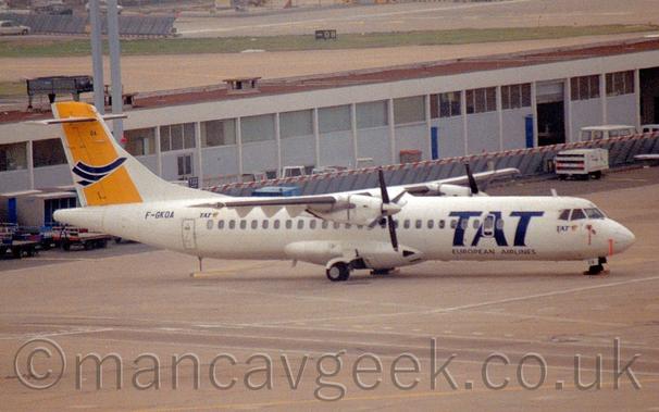 Side view of a high-winged, twin propellor-engined airliner parked facing to the right.
The plane is mostly white, with large, billboard-style blue "TAT" titles on the forward fuselage, with smaller blue "European Airlines" titles just below.
The tail has a diagonal yellow band overlaid a stylised image of a blue flying bird, with the blue registration "F-GKOA" on the upper rear fuselage just below.
Grey concrete apron fills the foreground, with a grey building with a flat red roof stretching across the background.