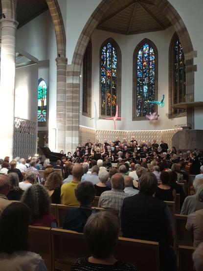 Vor drei Kirchenfenster und einem Rundbogen singt ein Chor. Im Vordergrund Publikum
