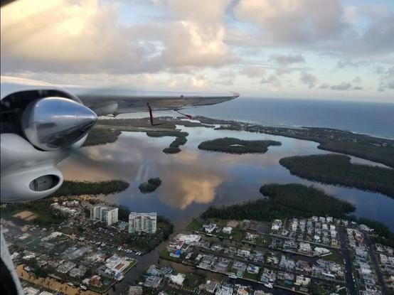 View from the window of an airplane, can see the left side propeller, houses, a lagoon, and the Atlantic Ocean.