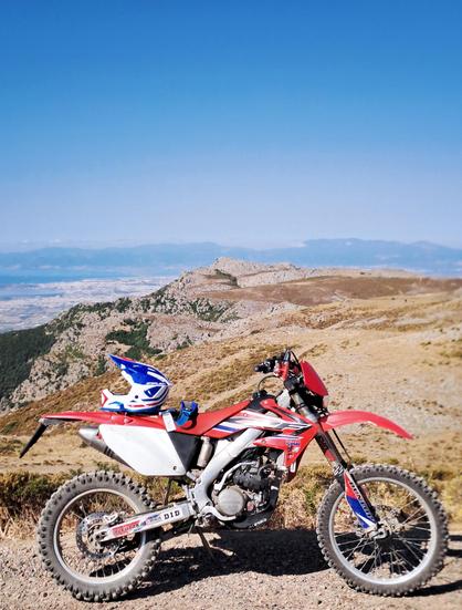 L'immagine mostra una motocicletta da cross rossa e bianca parcheggiata su un sentiero sterrato in un paesaggio montano. La motocicletta è dotata di pneumatici off-road e ha un casco blu e bianco posato sul sellino. Sullo sfondo, si estende un panorama mozzafiato con colline e montagne, e si intravede una città sottostante. Il cielo è azzurro e sereno, con poche nuvole. La motocicletta è un modello Honda CRF, come indicato dai loghi e dai colori.