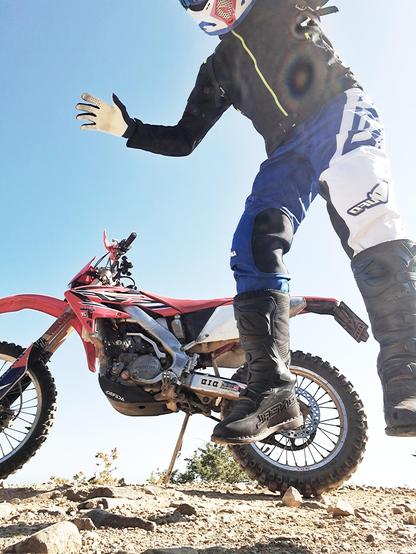 L'immagine mostra un motociclista in azione, in un contesto di motocross. Il motociclista è parzialmente visibile, con la parte superiore del corpo e le gambe in primo piano. Indossa un casco protettivo bianco con dettagli rossi e blu, guanti, una giacca nera con una cerniera gialla e pantaloni blu e bianchi con il logo "UFO" sulle gambe. I pantaloni sono realizzati in tessuto resistente, ideale per la protezione durante le attività di motocross. Indossa anche stivali neri robusti con la scritta "ONEAL" sul lato.

La motocicletta è una motocross rossa con dettagli neri e bianchi. È posizionata su un terreno roccioso e sabbioso, con un cielo azzurro e sereno sullo sfondo. La motocicletta è sostenuta da un supporto, indicando che il motociclista potrebbe essere in procinto di saltare o di eseguire una manovra. La scena trasmette un senso di azione e avventura, tipico delle attività di motocross.