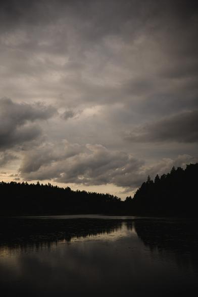 A Photograph of a forest lake at close to sunset. The bottom third show the lake and a hilly spruce and pine forest on the other side of the lake.

Above the treeline is a partly cloudy sky.

In the water are water lilies and the sky and trees are reflected in the almost calm water.