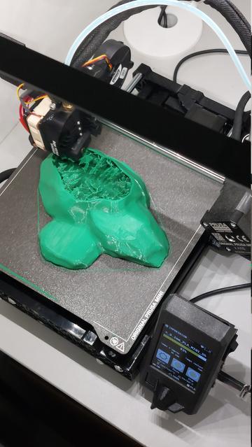Photo de la pièce en cours d'impression dans un plastique vert dans l'imprimante. Le haut n'est pas encore refermé et on voit les structures de remplissage en forme d'éclairs.