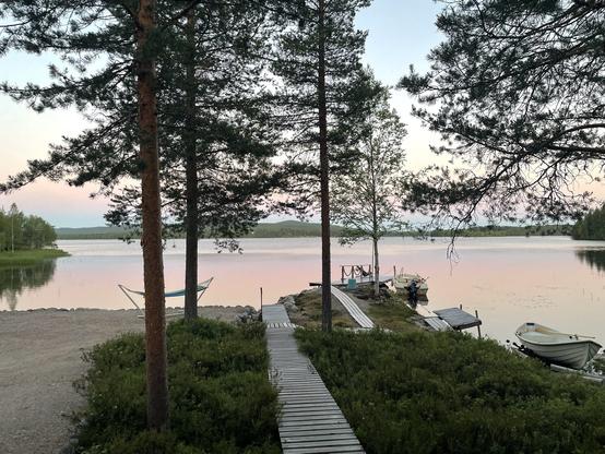 Maisema lappilaiselta järvenrannslta, hiekkaranta, suopursujen yli eräänlsiset pitkospuut johtavat lsiturille js järvelle. Vastarannalla kajastavat hahupuiden peittämät vaarat. Taivas ja järvi kajastavat vaaleansinistä ja -punaista valoa. 
Kello on 1 a.m.