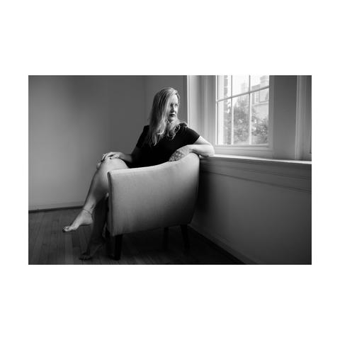 Portrait in black and white of a woman sitting in a sofa next a window and looking away to the outside world