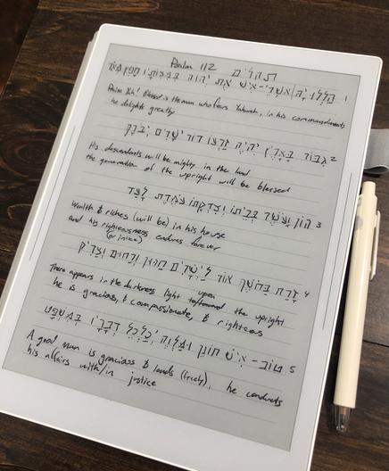 A white tablet sits on a wood surface. The black text on the table is the handwritten text of Psalm 112:1-5 written in Hebrew and then translated into English.