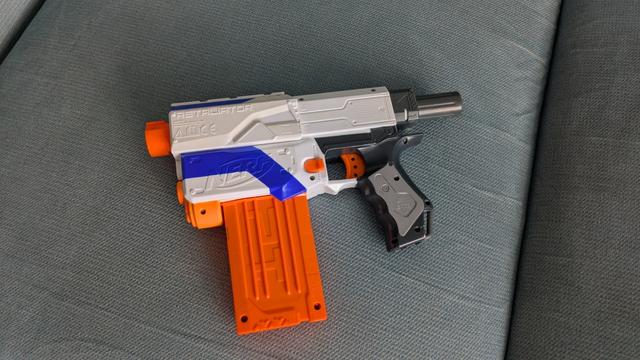 Fragment karabinu nerf retaliator, bez kolby i lufy. Większość jest z białego plastiku poza czarnym uchwytem, niebieskimi elementami dekoracyjnymi i kilkoma pomarańczowymi fragmentami. Przypomina to pistolet z masywnym, pomarańczowym magazynkiem.