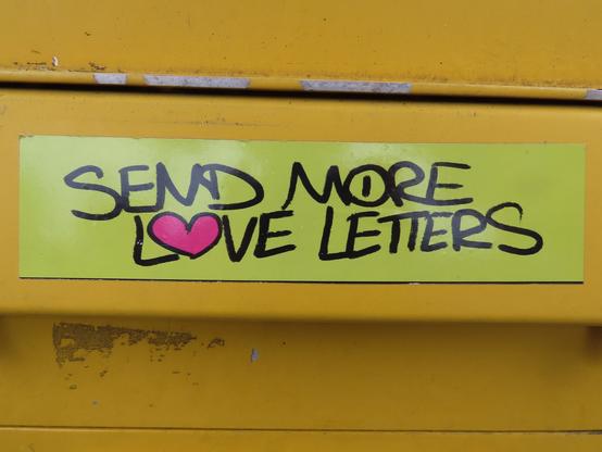 Grünlich-gelber Sticker auf der Klappe vor dem Einwurfschlitz eines gelben Briefkastens der Post. Auf ihm steht "SEND MORE LOVE LETTERS" in schwarz, wobei das O in LOVE wie ein Herz geformt und pink gefüllt ist.