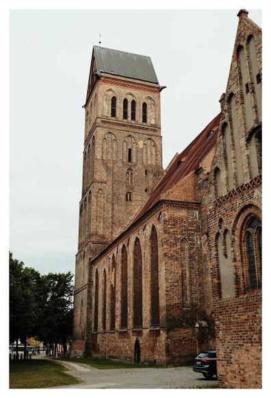 Backsteingotische Kirche mit hohem Turm und spitzem Dach. Die Fassade zeigt schlanke Spitzbogenfenster und dekorative Backsteinmuster. Ein Auto parkt an der Seite, Bäume und Kopfsteinpflaster rahmen das Bild.