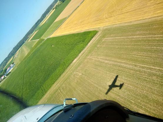 View from a light airplane across some green and yellow fields. Parts of the cowling and instrument panel are visible. The horizon is visible, canted in the upper left hand corner of the image. The shadow of the plane can be seen on the field in the lower right corner. A small grass landing strip is visible behind some fields on the horizon.