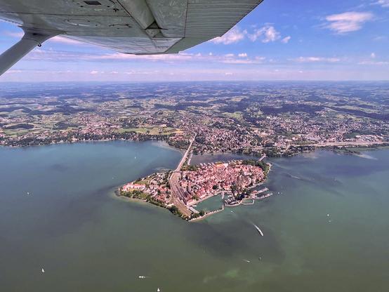 Aerial view of the town of Lindau, situated on an island in Lake Constance. The island is connected to the shore by two bridges, one of them carrying train tracks leading to a station on the island. The wing of the small airplane can be seen in the top of the picture. The sky is blue with some scattered clouds, and the lake reflects it in shades of turquoise. Several boats are visible on the surface.