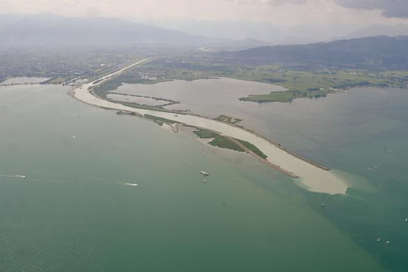 Aerial view of the river Rhine entering Lake Constance. The river is lead quite far into the lake by artificial embankments, and its colour is remarkably lighter than that of the lake. The sky is somewhat hazy.