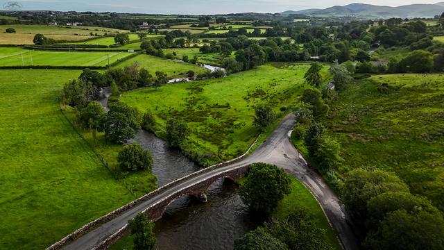 Aerial shot of narrow road bridge over River Ehen with farmland stretching into distant mountains