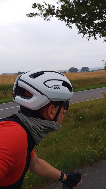Ein Radfahrer mit weißem Helm und orange-farbenen Trikot von der Seite auf einem Radweg parallel zu einer Landstraße.