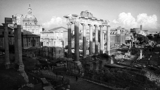 A black and white photograph of the Roman Forum in Rome. The image is dominated by the ruins of several large, fluted columns in the foreground, part of the Temple of Saturn. To the left, the dome of the Santi Luca e Martina church is visible, along with the facade of the Curia Julia. Further in the background, a path winds through more ancient ruins, with the arches of the Colosseum partially visible in the distance on the right. The sky is partly cloudy.