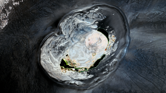 A satellite picture of a volcanic structure amidst black sand. The structure is clearer than its surroundings and we can see lakes and a crater in the middle of it.