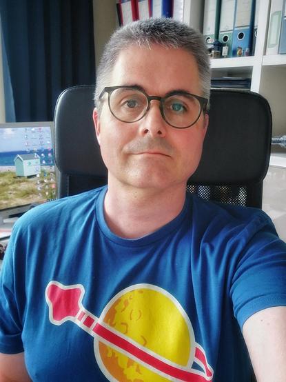 Ein Selfie von mir wie ich im Homeoffice an meinem Schreibtisch sitze. Im Hintergrund ist eine Ecke von meinem Bildschirm zu sehen.