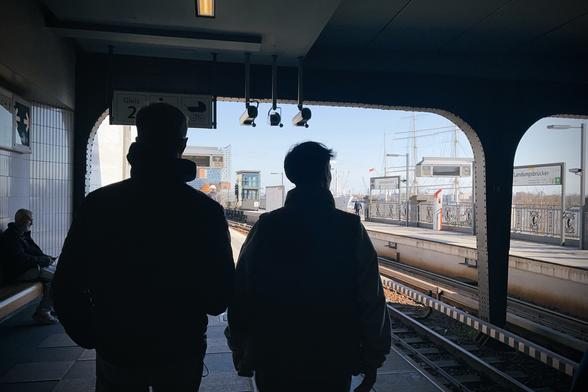 A chest-height photograph along the Landungsbrücken U-Bahn platform in Hamburg. The camera is located under a flat roof made from riveted steel. The area under the roof is dark, but visible. Bright sunshine outside that is "correctly" exposed. The main subject are two male-looking persons walking away from the camera side by side, visible from head to hips. Their silhouettes are framed by the station roof.
