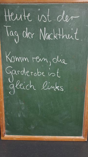 Auf einer grünen Tafel steht mit weißer Kreide geschrieben:
Heute ist der Tag der Nacktheit. Komm rein, die Garderobe ist gleich links.