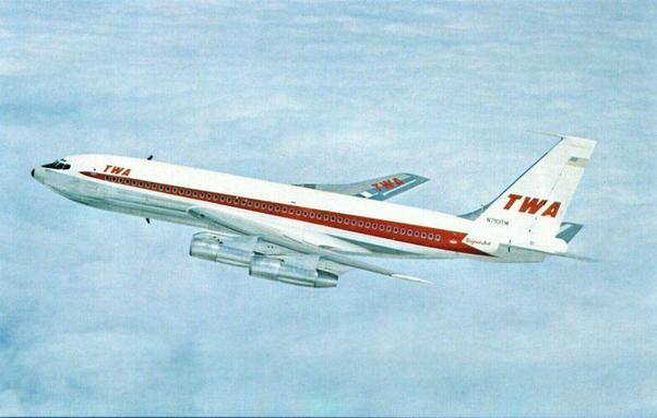 The Airplane: The main focus is a TWA (Trans World Airlines) Boeing 720, identifiable by its fuselage shape, engine placement, and the prominent TWA logo on the tail and forward fuselage. The airplane is predominantly white with a red stripe along the side, featuring the TWA logo in red lettering. The registration number "N793TW" is visible on the tail. The word "SuperJet" is also visible near the rear of the plane.

    The Setting: The airplane is shown in flight against a backdrop of a light blue, slightly cloudy sky. The clouds are relatively thin and wispy, allowing the plane to be clearly visible.

    The Image Quality: The image appears to be a scan or reproduction of a photograph, possibly from a postcard or brochure, given the slightly aged and somewhat faded look. The colors are muted and slightly soft.