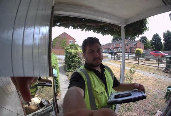 A bearded young man rings a video doorbell to make an unsolicited sales approach. He wears a yellow hi-vis waistcoat and carries a thick clipboard with a smartphone laid on it.  He claims to be an “electrical surveyor” but is just trying to get householders to sign up his company to obtain wayleaves for supply cables on their property.