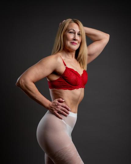 Woman with blonde hair models in a red lace bra and sheer white tights. Her right hand rests on her waist and her left arm is raised, touching her head. The background is a plain, dark gradient. She wears a ring on her left hand.