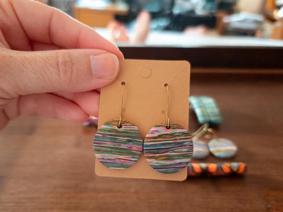 In the blurred background, all of the finished pieces, except for one pair of earrings. Those are being held on an earring card by me in the foreground. They are about .75-inch polymer clay circles hanging from brass ear wires. The design is a messy stripe with a variety of pastel colors, and even a bit of sparkle. They have been sanded to be exceptionally smooth, and buffed to a nice shine.