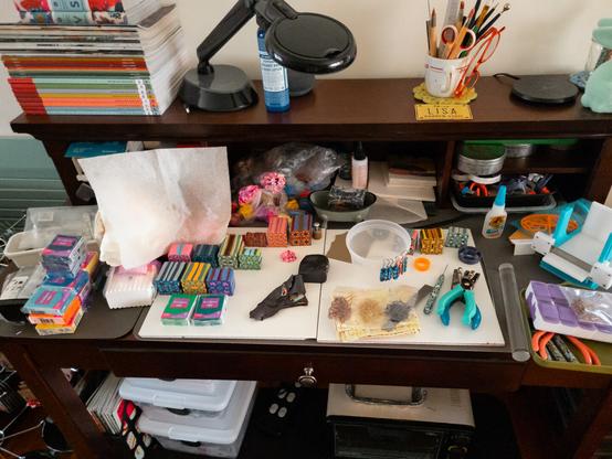 A messy desk, with unfinished polymer clay projects, finished cane designs, finished jewelry, jewelry-making supplies and tools, and other unrelated stuff scattered about.