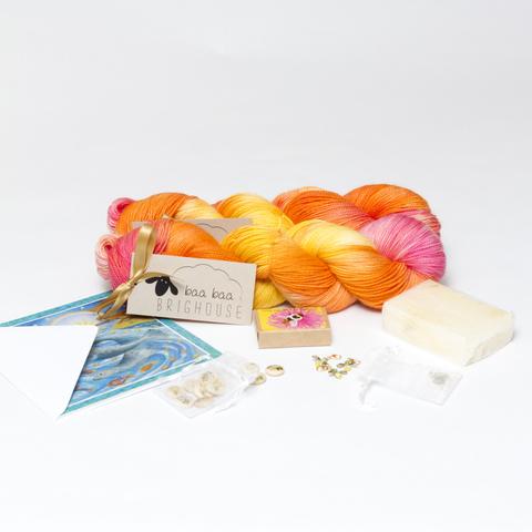 Image shows two skeins of yarn, one behind the other, in shades of orange, yellow and pink. In front, from left to right, is a card, a bag of buttons, a box of seed balls, a bag of stitch markers and a bar of soap.