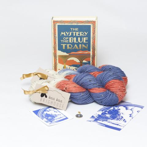 Picture shows two skeins of yarn in shades of blue, orange and white, positioned one behind the other. Behind the yarn, stood on its end, is a hardback copy of The Mystery of the Blue Train. In front of the yarn, from left to right, is a sticker, a stitch marker and a postcard.