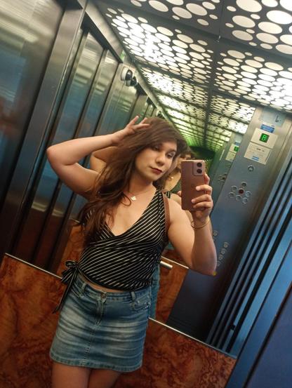 Mirror selfie of a brunette with long hair, wearing dark red lipstick and a black with white diagonal stripes top with mid deep cleavage in V. She also wears a short jeans skirt. She poses with her free hand slightly touching her hair at the scalp.