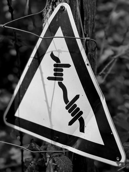 View of a ‘caution barbed wire’ sign on a fence post. (Version françæ) Vue d’æn panneau 'attention barbelés' apposæ sur un piquet de clôture.