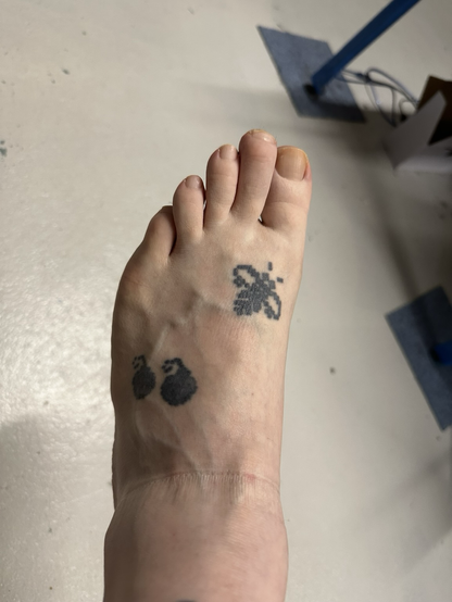A closeup of my bare left foot, showing a tattoo of two pixelated bombs and a bee.