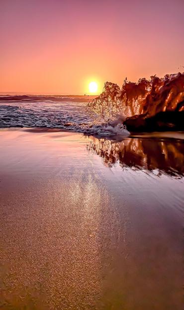 Sunset over the ocean with a bright, orange sun near the horizon. A wave breaks over a rock on the right, creating splashes. The wet sandy beach reflects the colorful sky, transitioning from pink to orange.