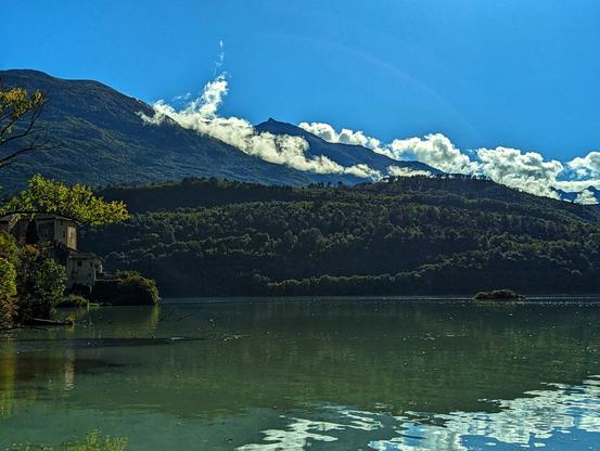 A scenic shot on a sunny day shows a calm lake reflecting the sky and clouds, with a small, tree-covered islet towards the right. In the background, lush green mountains rise, their peaks partially obscured by white clouds. On the left side of the lake, a stone building with a red roof is nestled amongst trees, with more buildings barely visible behind it. The sky is bright blue with scattered clouds.